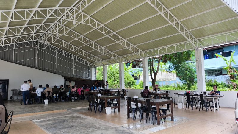 ภายในโรงเรือนโล่ง โต๊ะไม้ตัวยาวเรียงใต้โครงหลังคาเหล็กสีขาว มองเห็นต้นไม้ด้านนอก