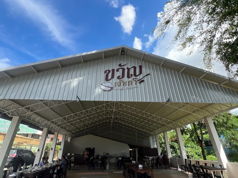 โรงเรือนโล่งหลังคาเหล็กสูงของร้าน มีป้ายชื่อขวัญเจ้าเก่าบนหน้าจั่ว โต๊ะเก้าอี้เรียงอยู่ด้านใน ใต้ฟ้าสีคราม